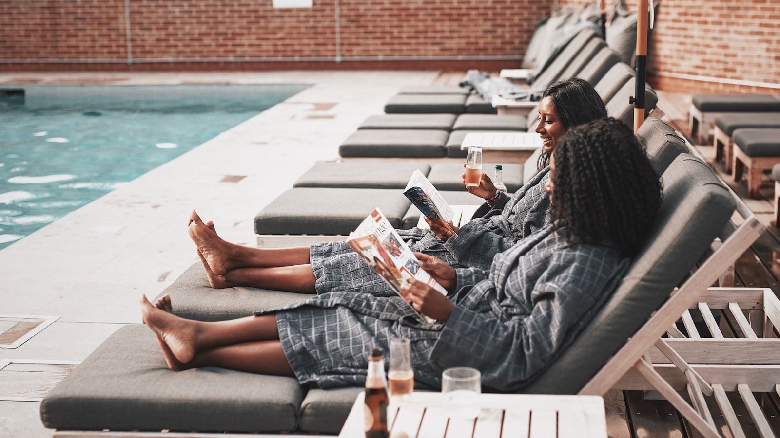 Two women laying on sunbeds outside next to a swimming pool drinking prosecco and reading magazines in robes at the Talbooth House and Spa 